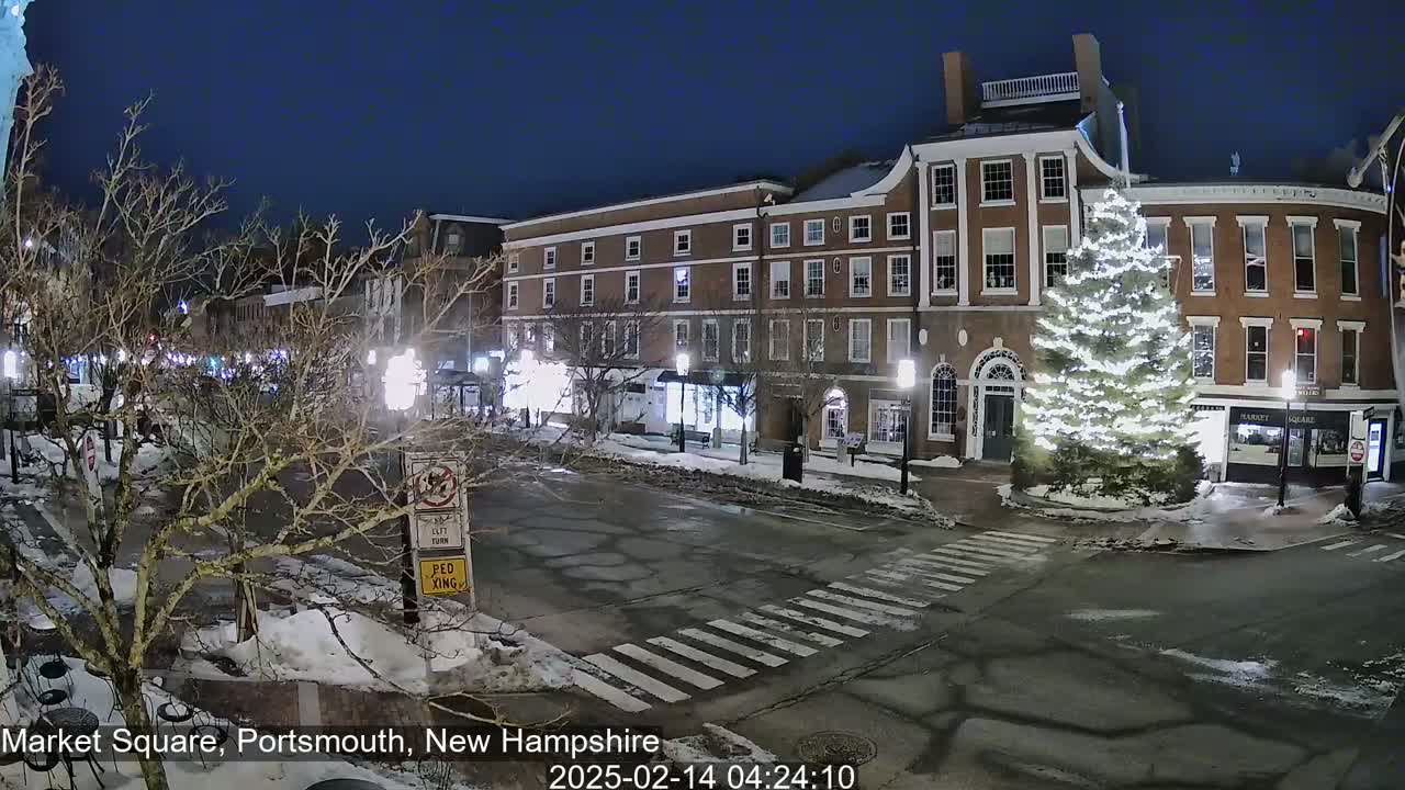 A snow-covered town square at night, illuminated by streetlights and a large, brightly lit Christmas tree in front of brick buildings.