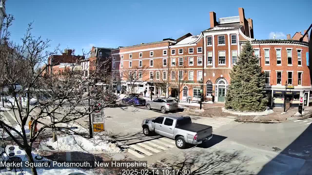 A sunny day in a brick-lined town square shows a few cars and pedestrians, with patches of snow and bare trees on the ground.