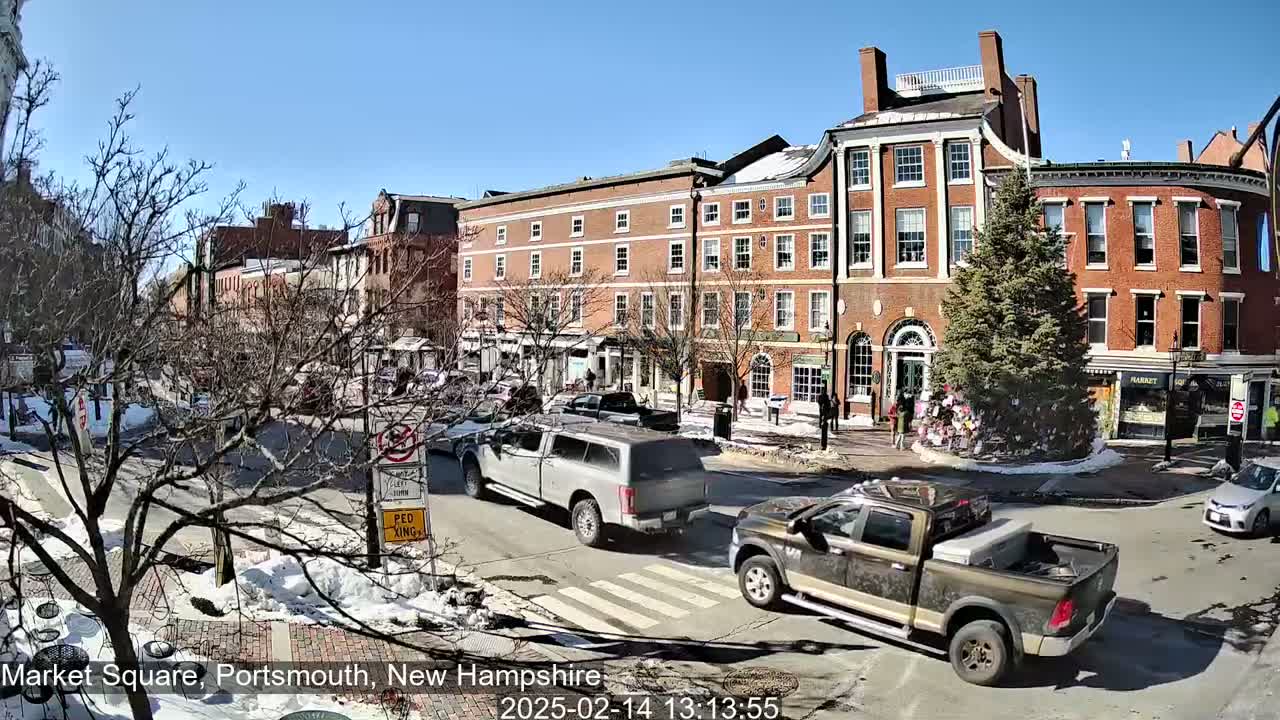 A sunny day in a town square shows several brick buildings, a few cars driving on the street, and patches of snow on the ground.