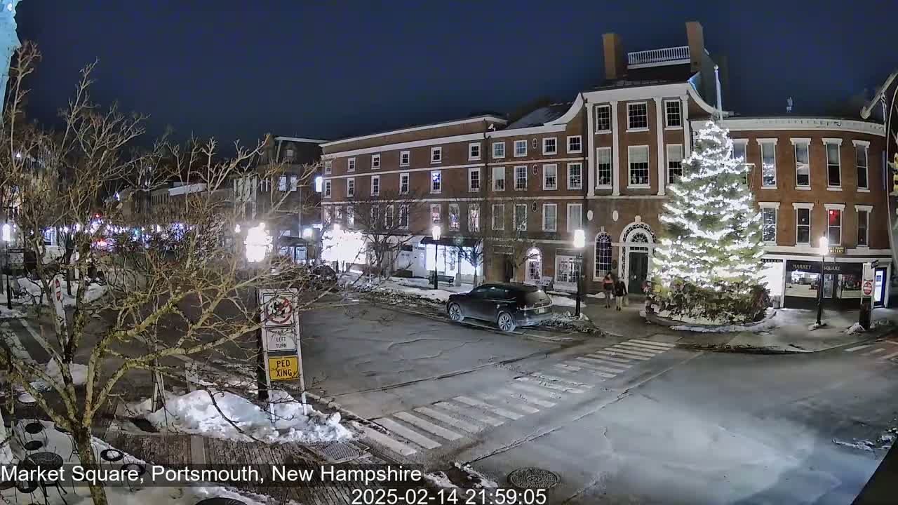 A snow-covered, nighttime street scene shows a town square with a large, illuminated Christmas tree in front of brick buildings.