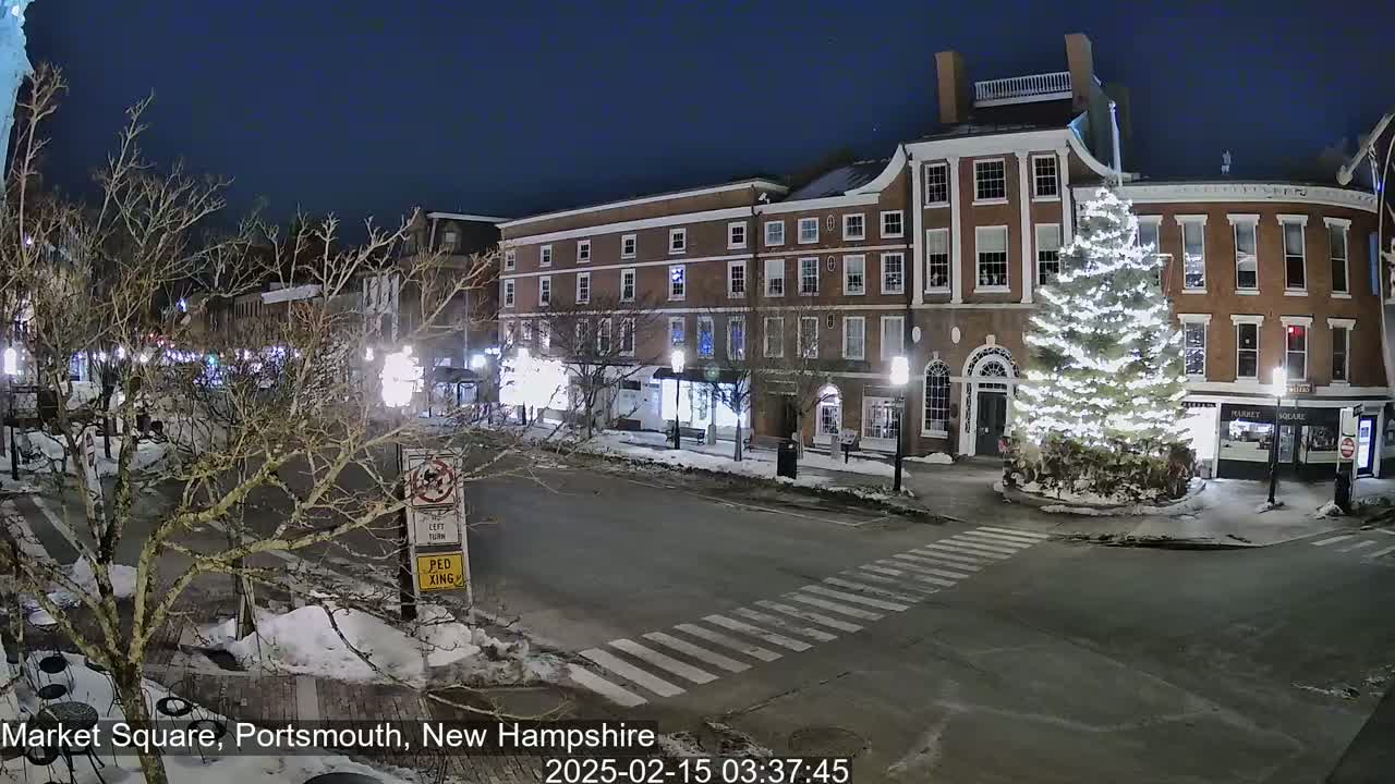 A snow-covered town square at night, illuminated by streetlights, features a large, decorated Christmas tree in the center and brick buildings lining the streets.