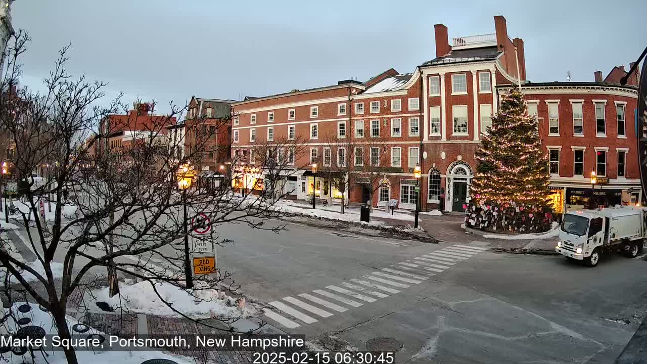 A snow-covered town square with brick buildings, a large decorated Christmas tree, and a garbage truck.