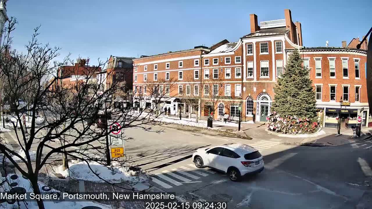 A sunny day in a town square shows several brick buildings, a decorated evergreen tree in the center of the square, and a white SUV driving across a crosswalk.