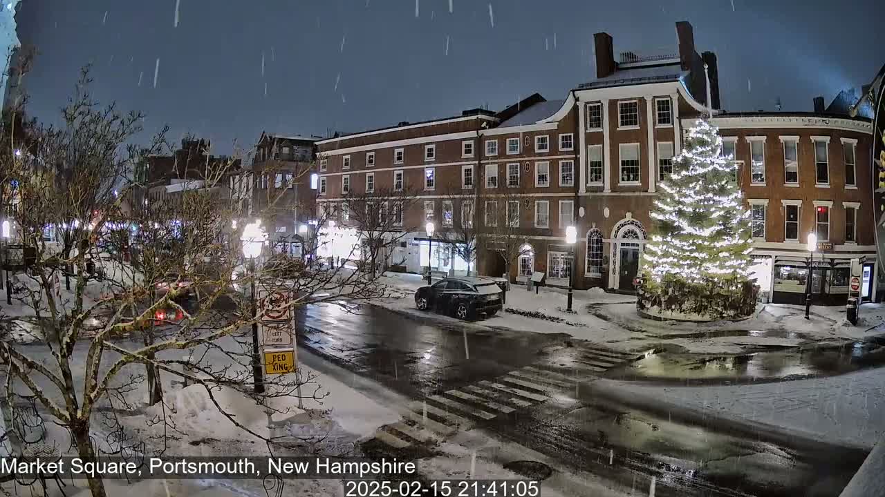 A snow-covered town square at night features a large illuminated Christmas tree in the center, surrounded by brick buildings and sparsely parked cars under falling snow.