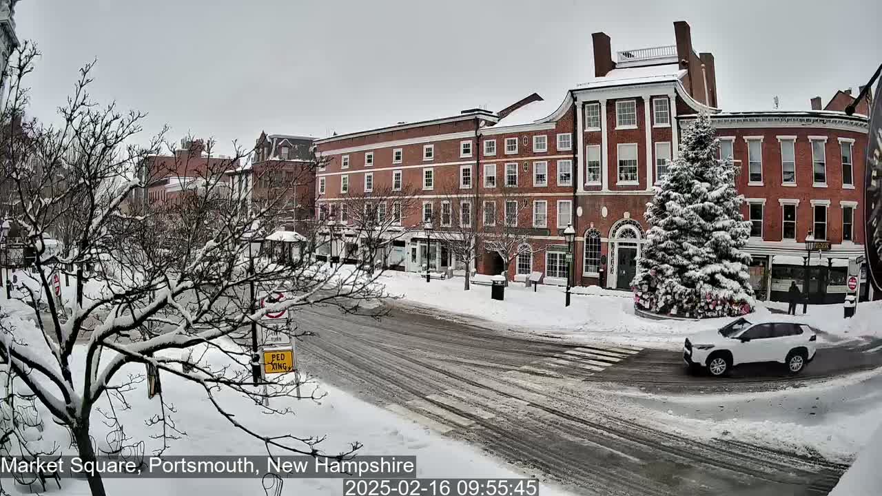 A snow-covered town square features several brick buildings, a snow-laden evergreen tree, and a white SUV driving on a slushy road.