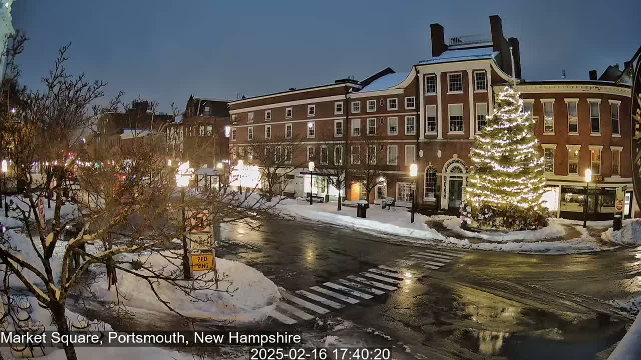 A snow-covered town square at night, featuring a large illuminated Christmas tree in front of brick buildings, with wet streets and some traffic.
