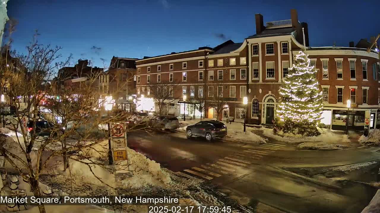 A snow-covered town square at night, illuminated by streetlights and a large, brightly lit Christmas tree, with cars driving on the streets.