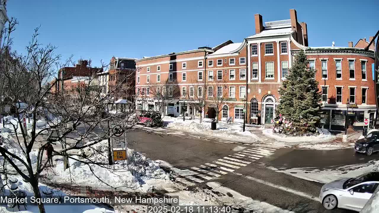 A sunny winter day in a snow-covered town square shows several brick buildings, a pedestrian crossing, and cars parked and driving.