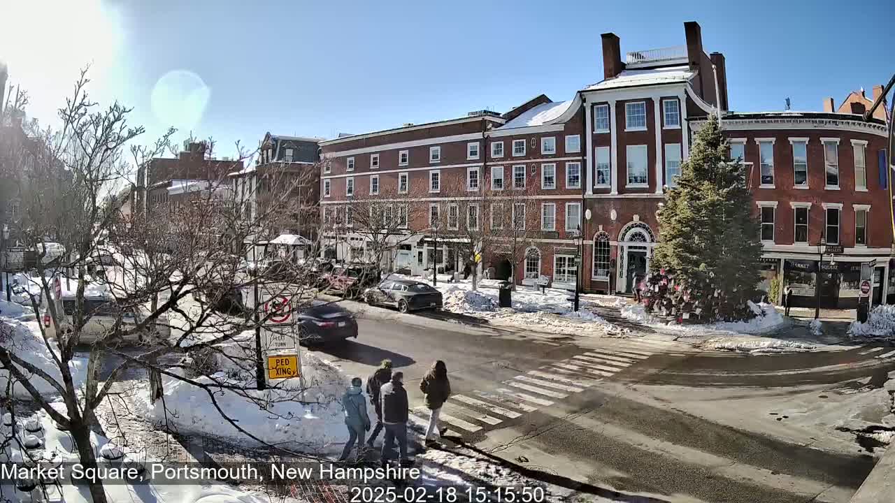 A sunny winter day in a town square with brick buildings, snow on the ground and sidewalks, and pedestrians crossing a street.