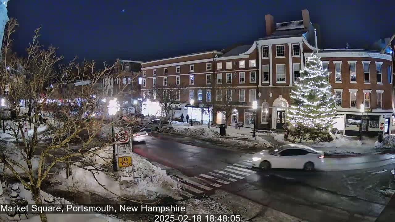 A snow-covered town square at night, illuminated by streetlights, features a large, lit Christmas tree in front of brick buildings and a white car driving through the square.