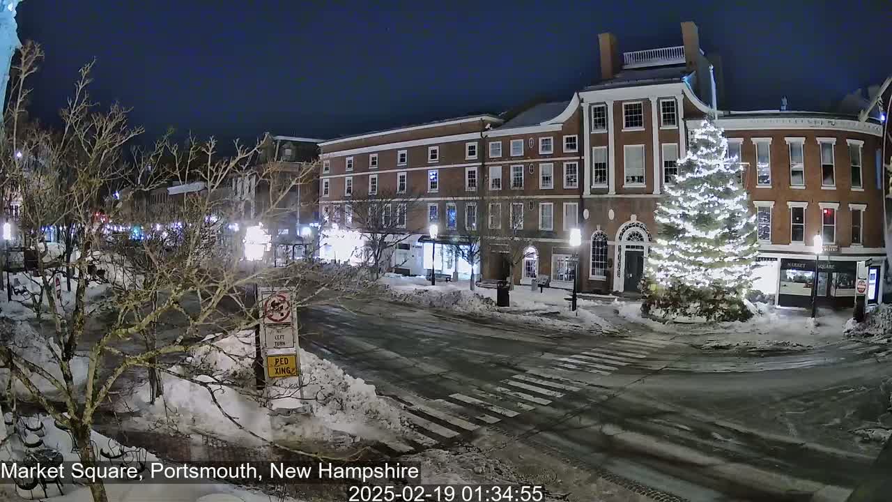A snow-covered town square at night, illuminated by streetlights, features a large, decorated Christmas tree in front of brick buildings.