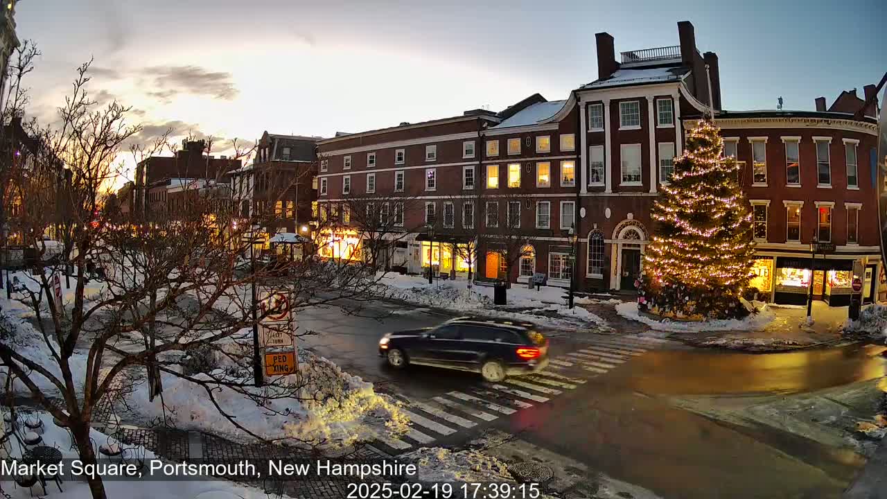 A snow-covered town square at twilight features a large illuminated Christmas tree situated between brick buildings, with a car crossing a pedestrian crossing.