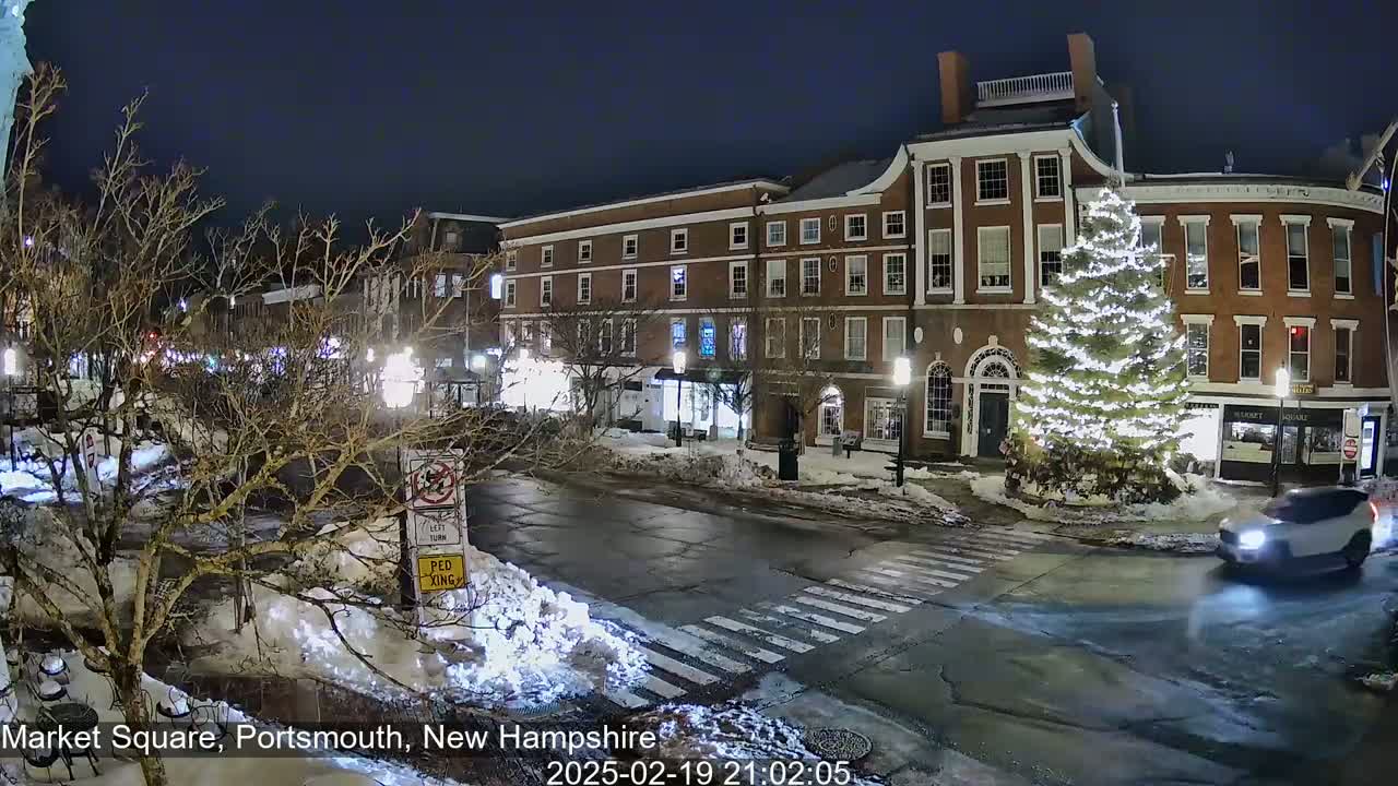 A snow-covered town square at night, illuminated by streetlights, features a large decorated Christmas tree in front of brick buildings.