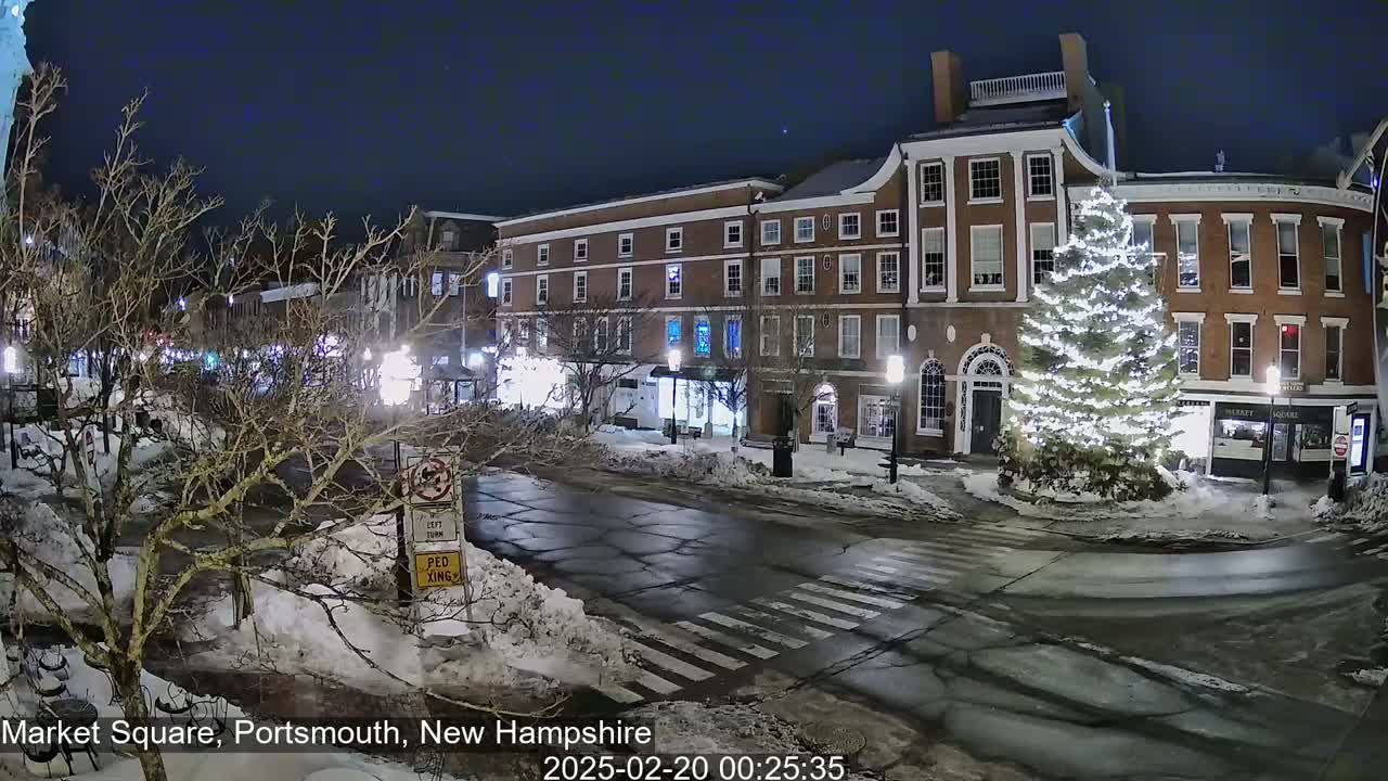 A snow-covered town square at night features a large, illuminated Christmas tree in front of brick buildings under a dark, clear sky.