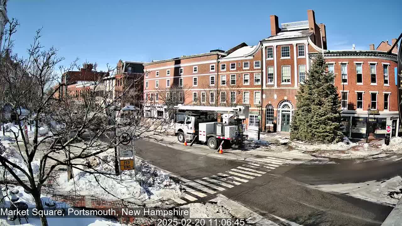 A sunny day in a snow-covered town square shows brick buildings, bare trees, a utility truck, and a crosswalk.