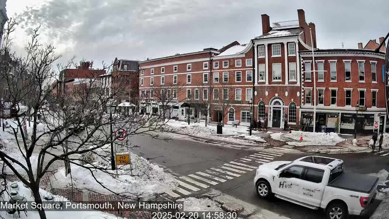 A snow-covered street scene shows several brick buildings under a cloudy sky, with a white pickup truck driving through a crosswalk.