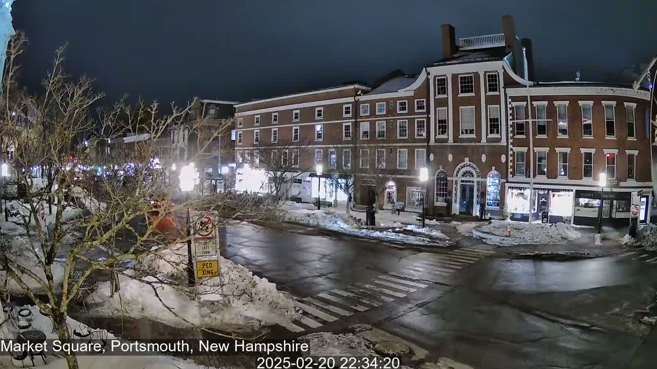 A snow-covered town square at night, with brick buildings illuminated, and a few cars visible on the wet street.