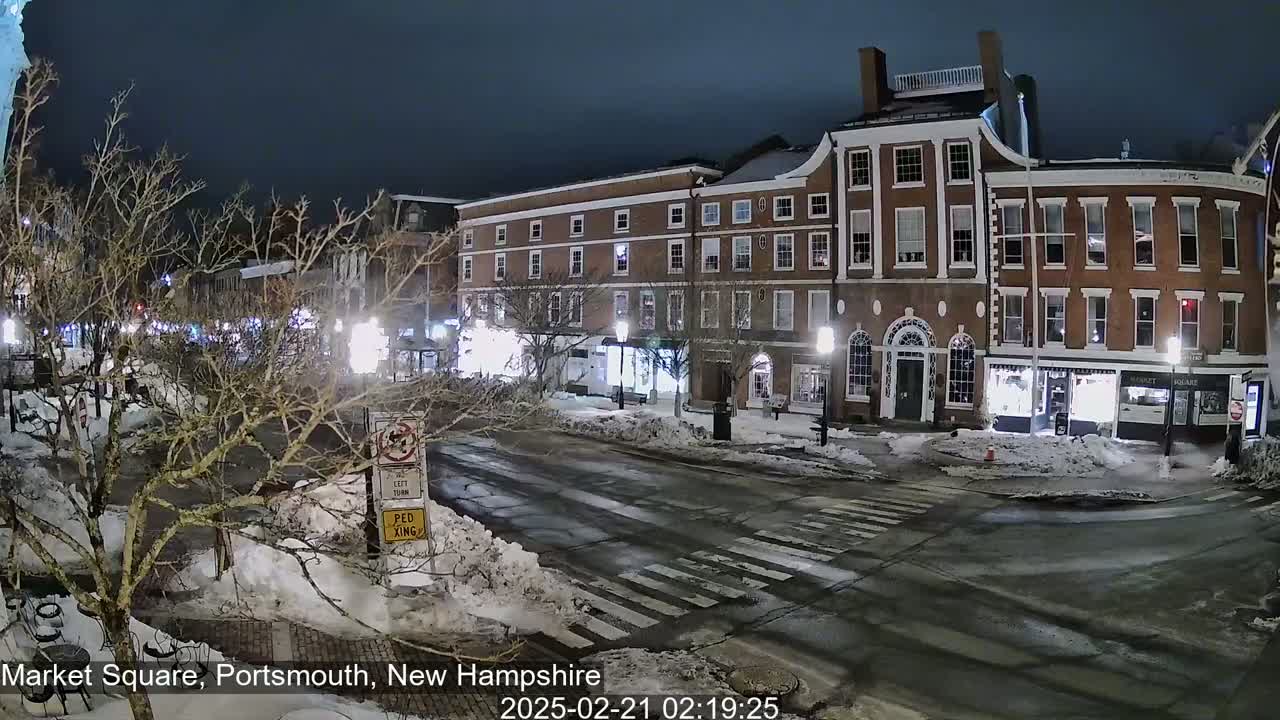 A snow-covered street at night, lined with brick buildings and streetlights, shows a mostly empty town square.