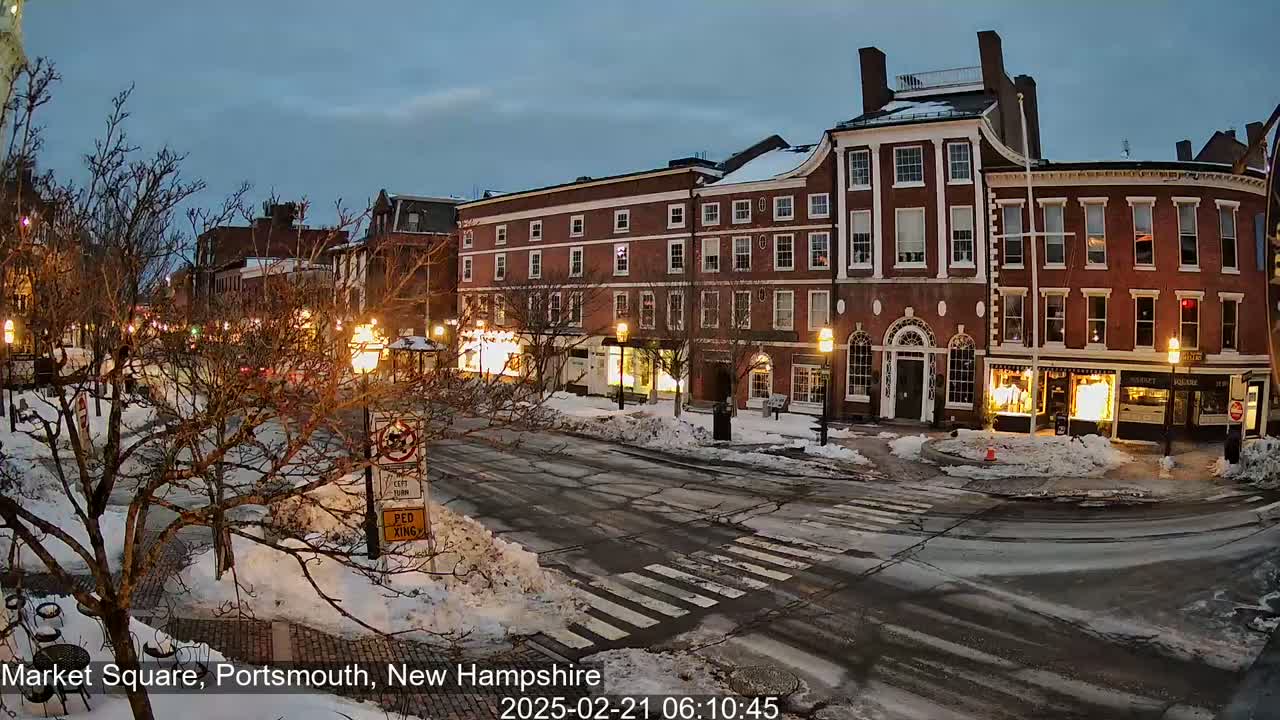 A snow-covered town square at twilight, featuring brick buildings with illuminated storefronts and bare trees lining the street.