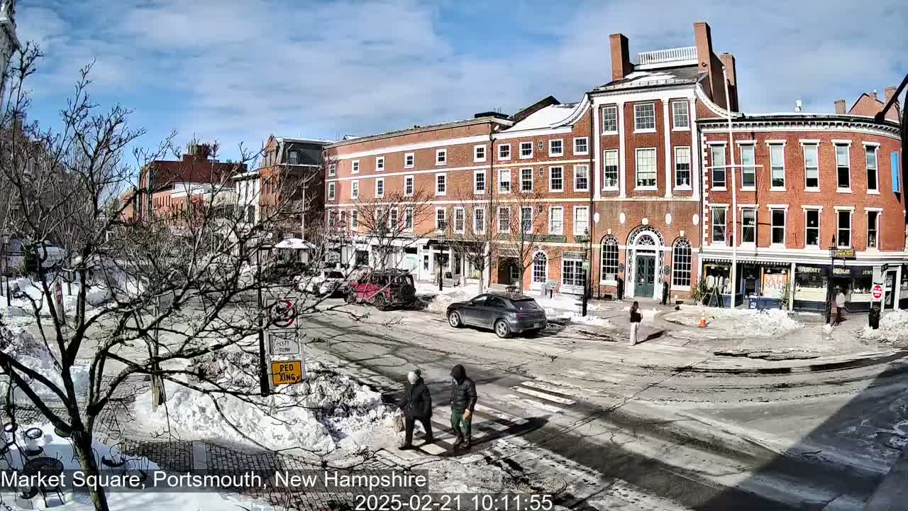 A sunny winter day reveals a snow-covered town square with brick buildings, parked cars, and pedestrians crossing the street.