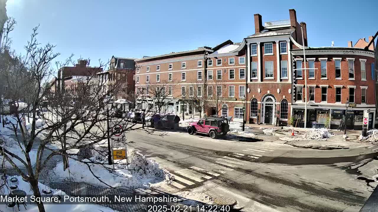 A sunny day in a city square shows several brick buildings, some with snow on the ground and parked cars.