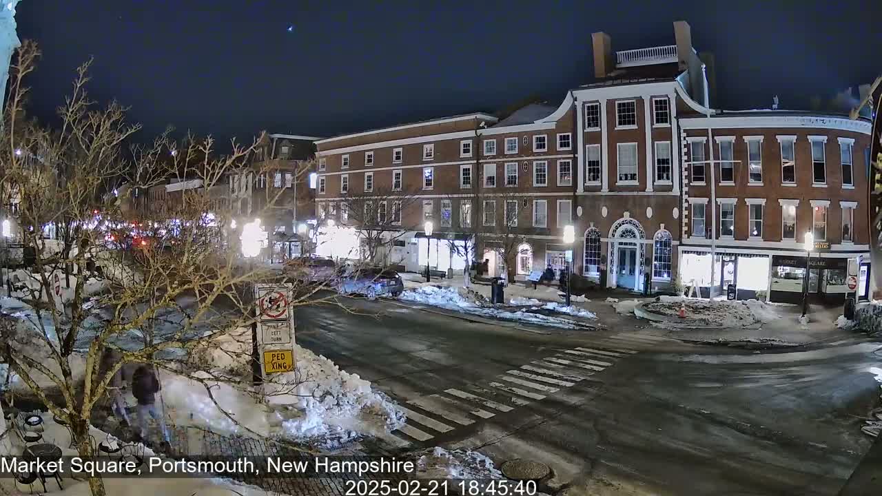 A snow-covered town square at night, with brick buildings lit up, a pedestrian crossing, and a few cars and pedestrians.
