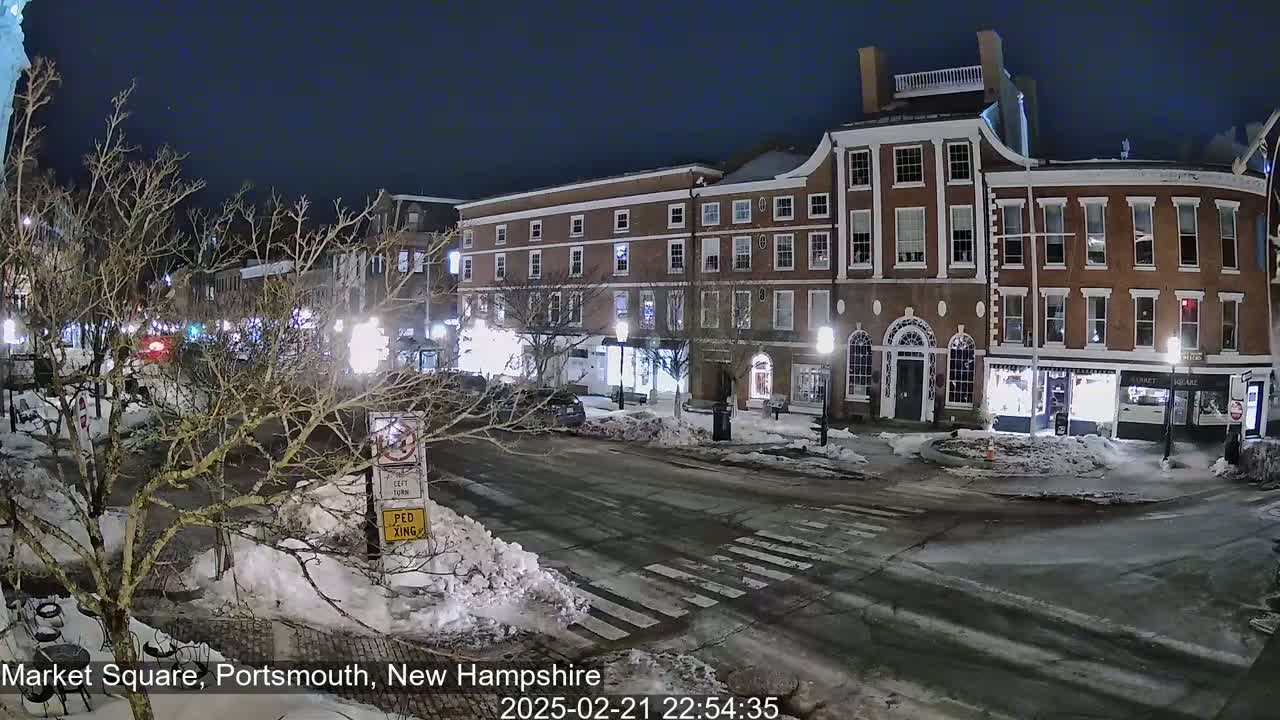 A snow-covered town square at night, illuminated by streetlights, shows several brick buildings and bare trees.