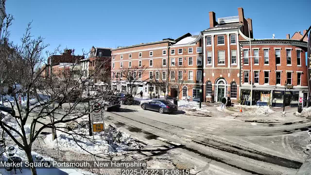A sunny day in a snow-dusted town square shows several brick buildings, cars, and a few pedestrians.