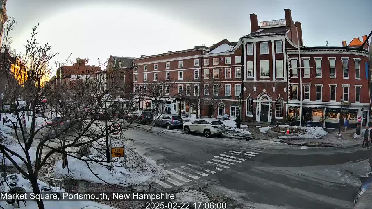 A snow-covered street scene shows several brick buildings and cars in a town square at dusk.