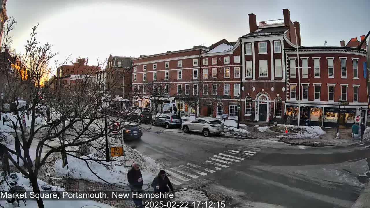 A snow-covered street scene shows several brick buildings at sunset with pedestrians and cars in a town square.