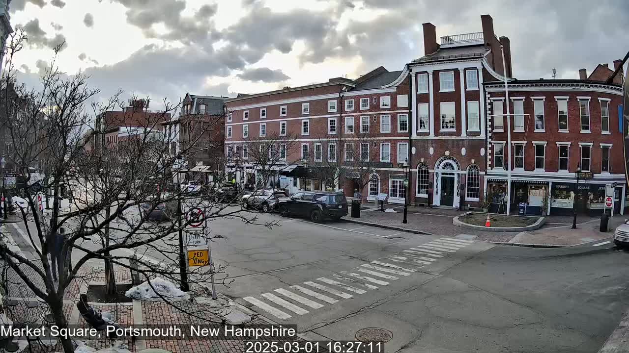 A partly cloudy day reveals a brick-lined town square with parked cars and sparse pedestrian traffic.