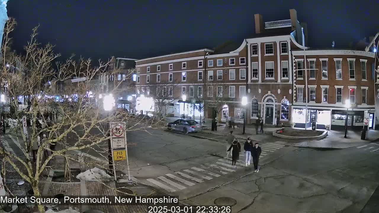 At night, a mostly empty town square with several pedestrians crossing a street in front of brick buildings.