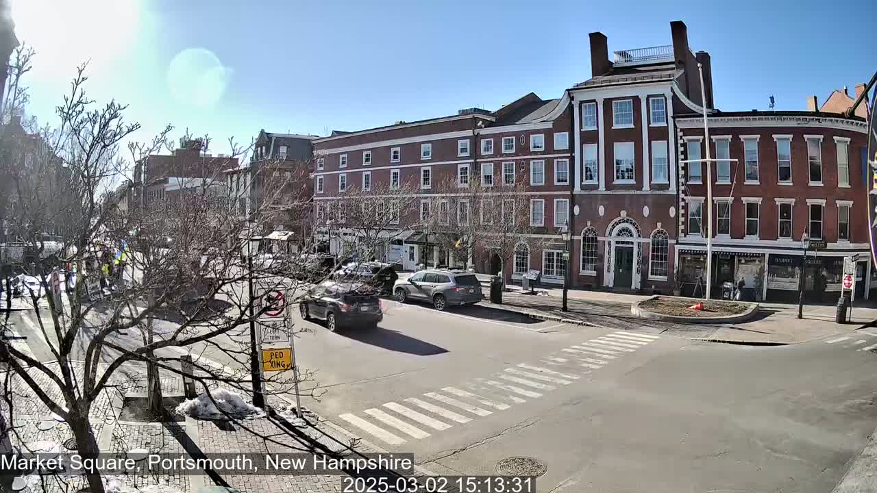 A sunny day in a city square shows brick buildings, bare trees, a crosswalk, and several cars driving on the street.