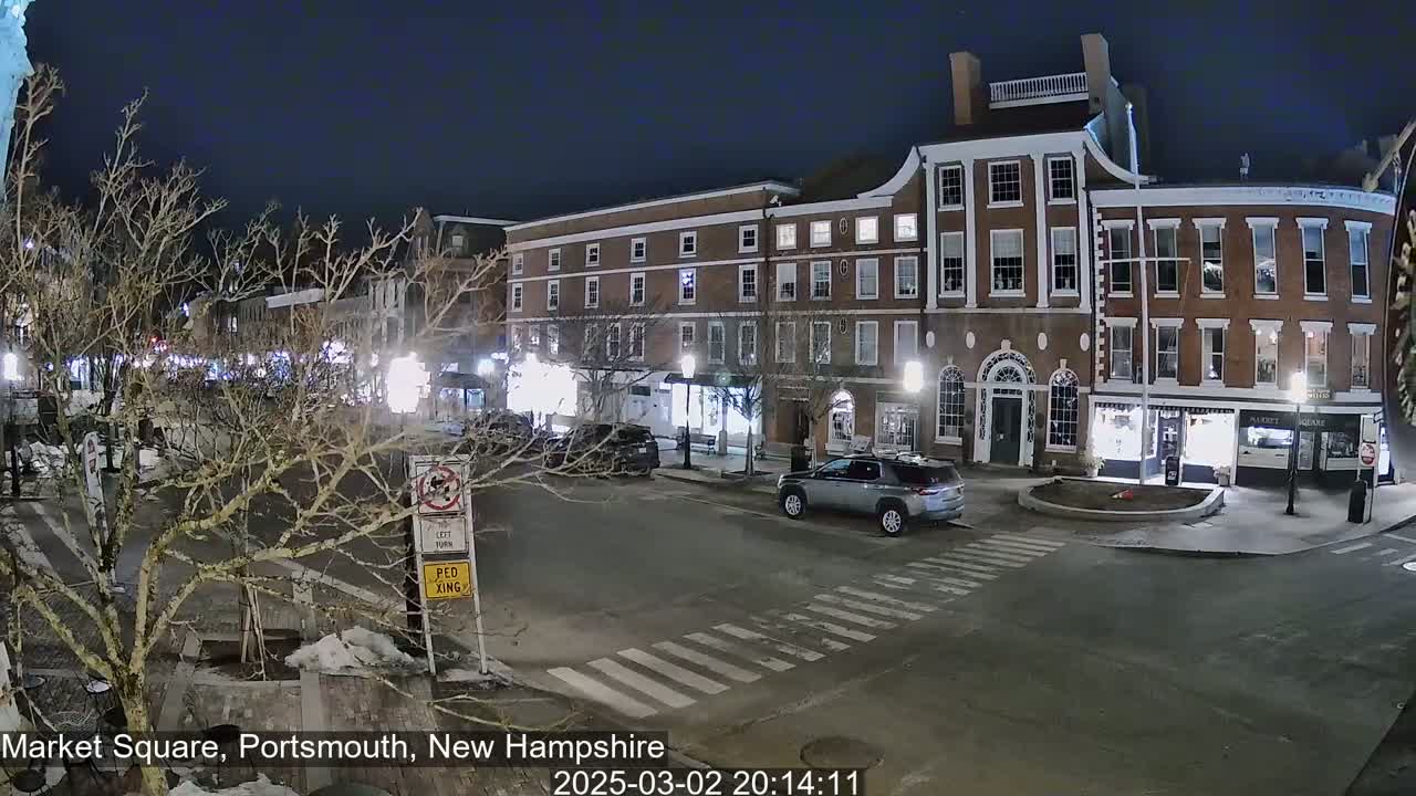 A nighttime view of a brick building-lined square with a few cars and bare trees under a dark sky.
