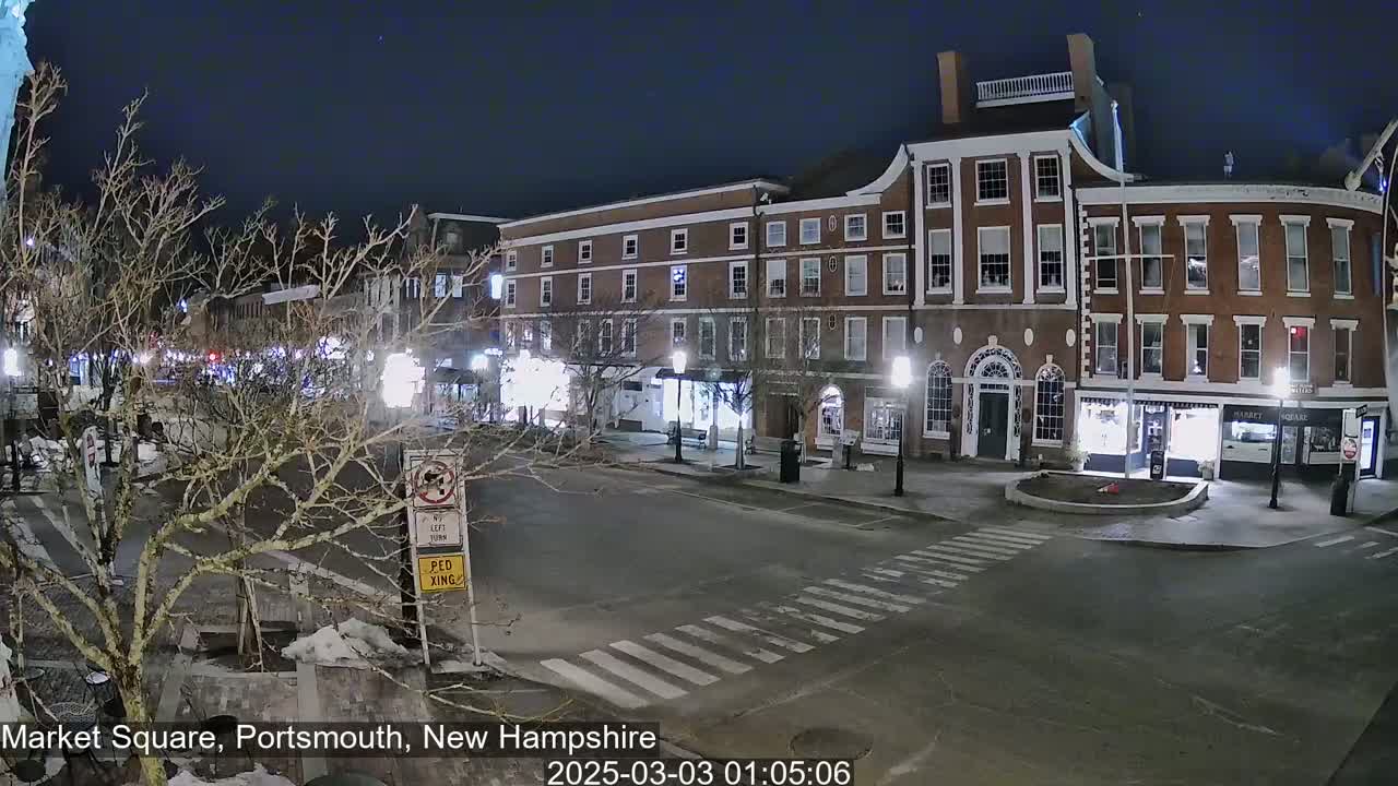A nighttime view of a mostly empty town square with brick buildings and bare trees under a clear, dark sky.
