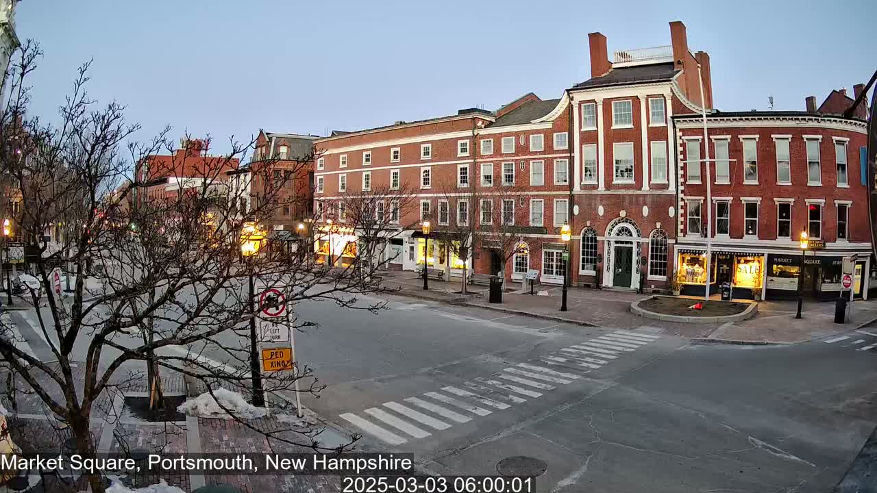 A mostly empty town square at dawn, featuring brick buildings with shops on the ground floor, under a clear, twilight sky.
