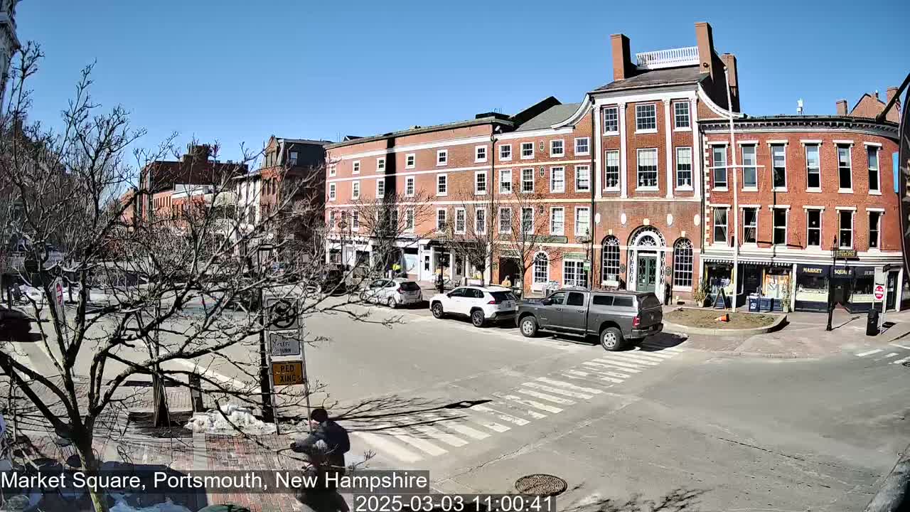 A sunny day in a town square shows several brick buildings, sparse, bare trees, and a few cars and a person crossing the street.
