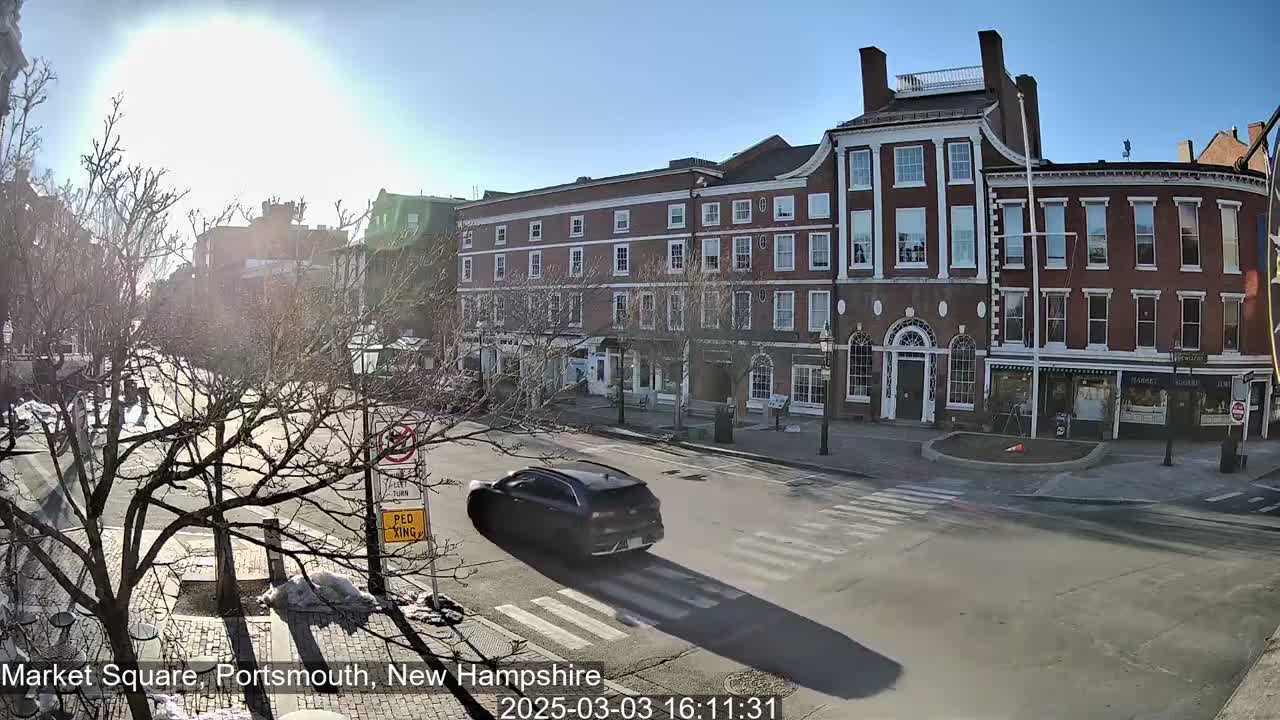 A sunny day reveals a city street scene with brick buildings, bare trees, and a dark-colored SUV driving past a crosswalk.