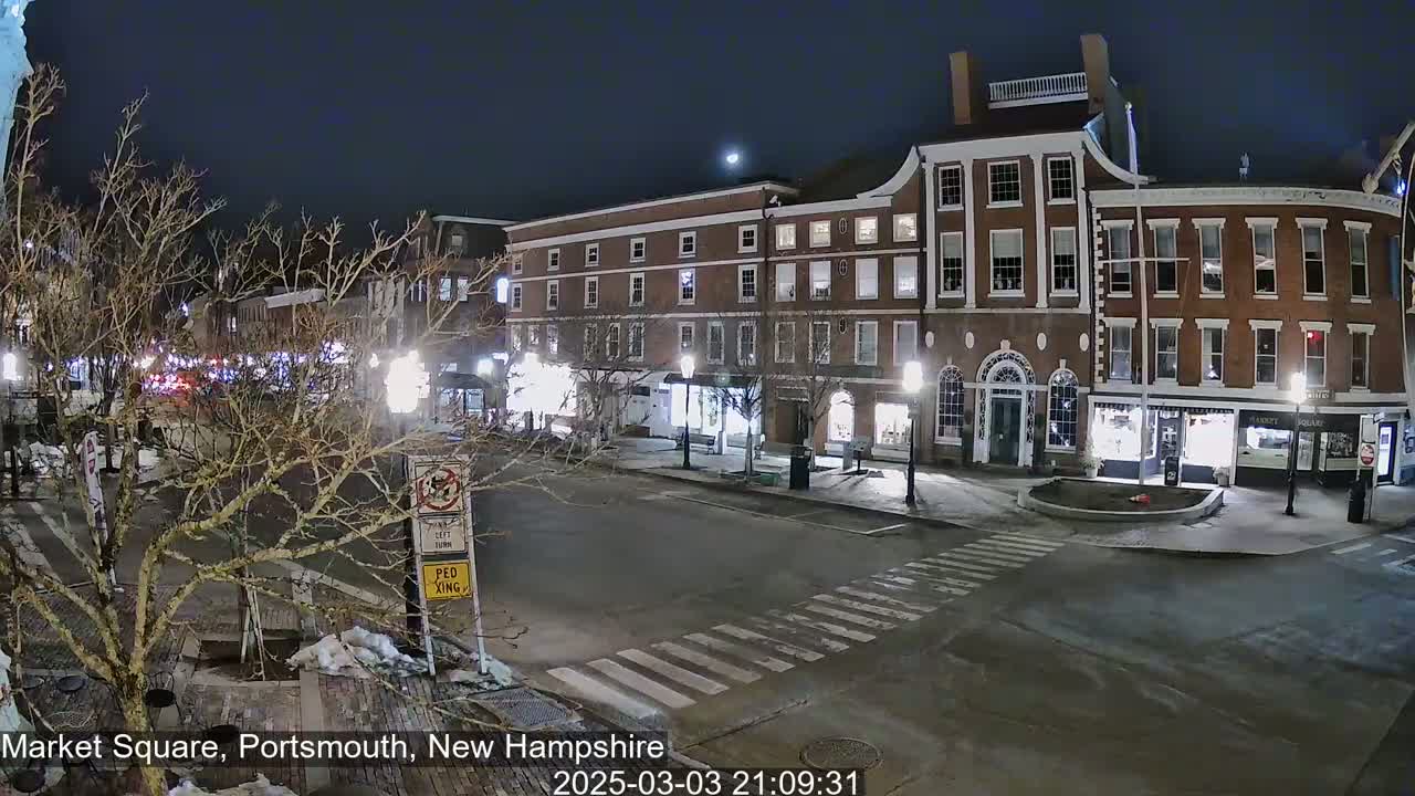 A nighttime view of a snow-dusted town square with brick buildings and streetlights, under a clear sky.