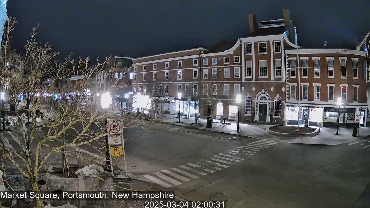 A nighttime view of a brick-lined town square with streetlights illuminating the empty street and surrounding buildings under a dark sky.