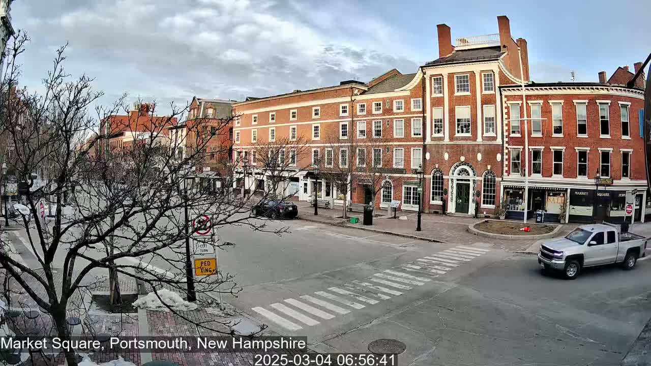 A mostly cloudy day reveals a city square with brick buildings, bare trees, and a silver pickup truck driving through a crosswalk.