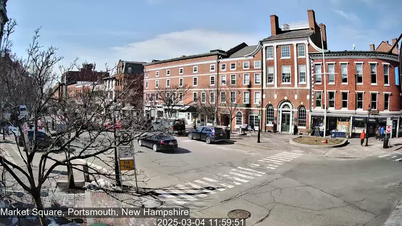 A sunny day in a city square shows several brick buildings, bare trees, and a few cars driving through a crosswalk.