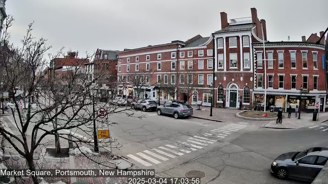 A mostly overcast day shows a brick-lined town square with several cars and pedestrians, some bare trees, and a crosswalk.