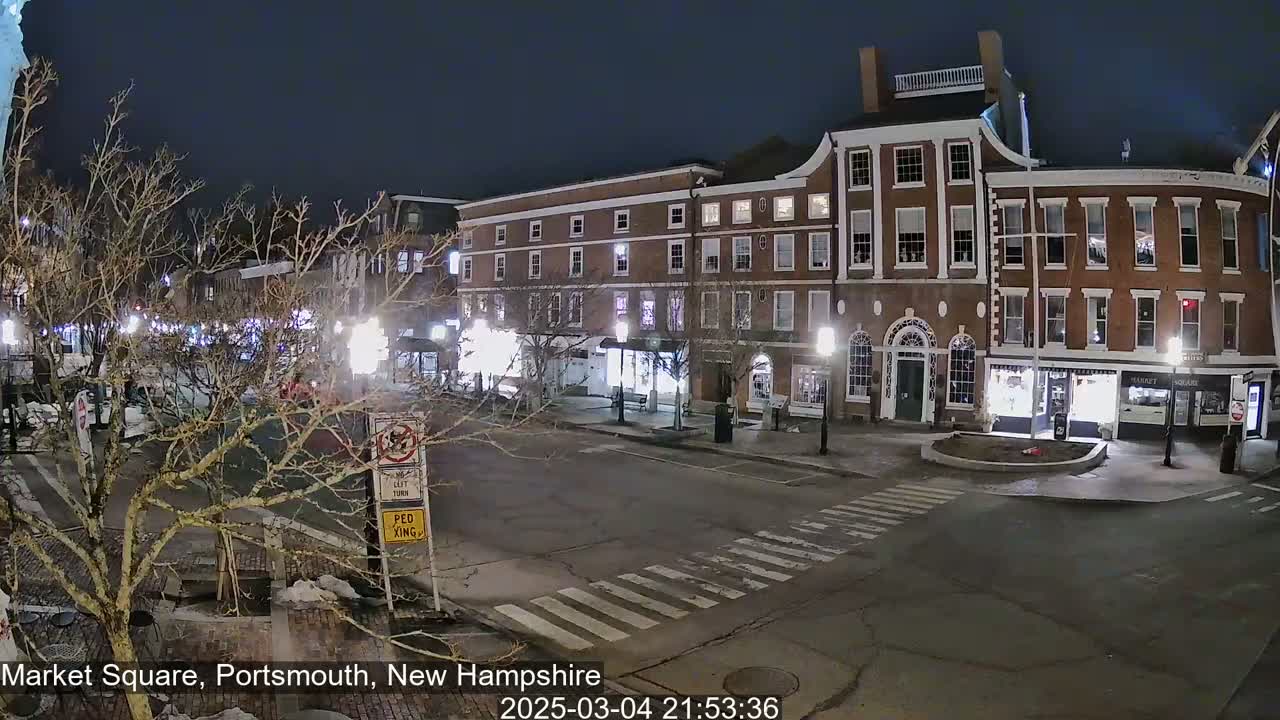 A nighttime, city street scene shows a mostly empty, brick-lined square with a few parked cars and bare trees under dark skies.