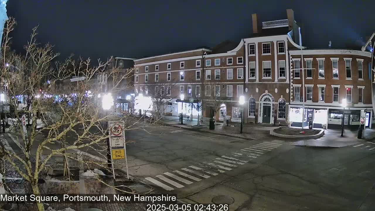 A nighttime view of a mostly empty town square with brick buildings, bare trees, and streetlights.
