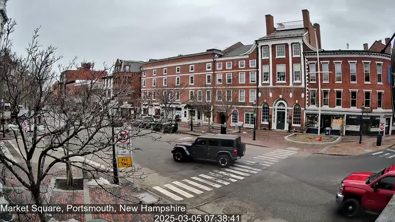 A gray Jeep and a red truck are driving through a city square on an overcast day, past several brick buildings.