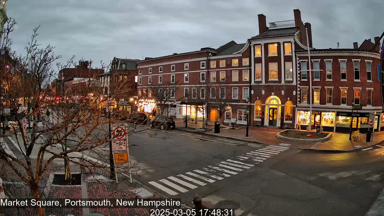 A city square at twilight, under a cloudy sky, features several brick buildings with illuminated windows and a pedestrian crossing in the foreground.