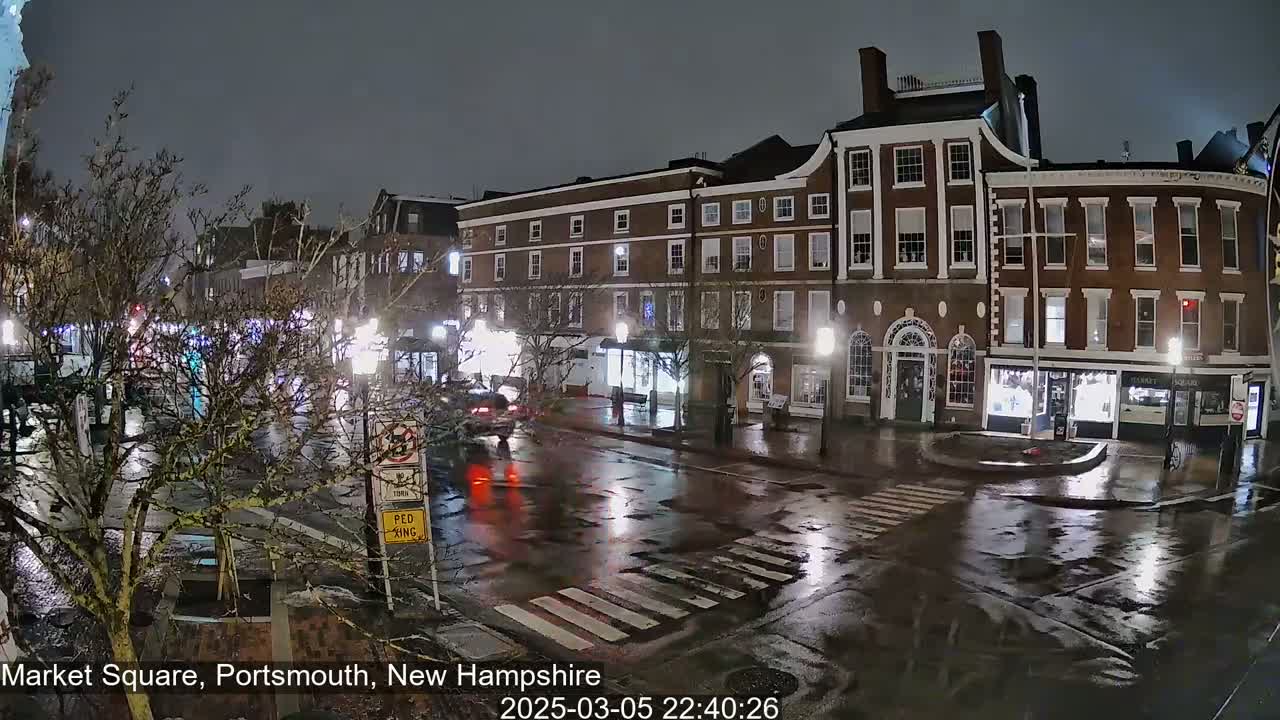 A rain-slicked town square at night, showing several multi-story brick buildings with streetlights illuminating the wet pavement and sparse traffic.