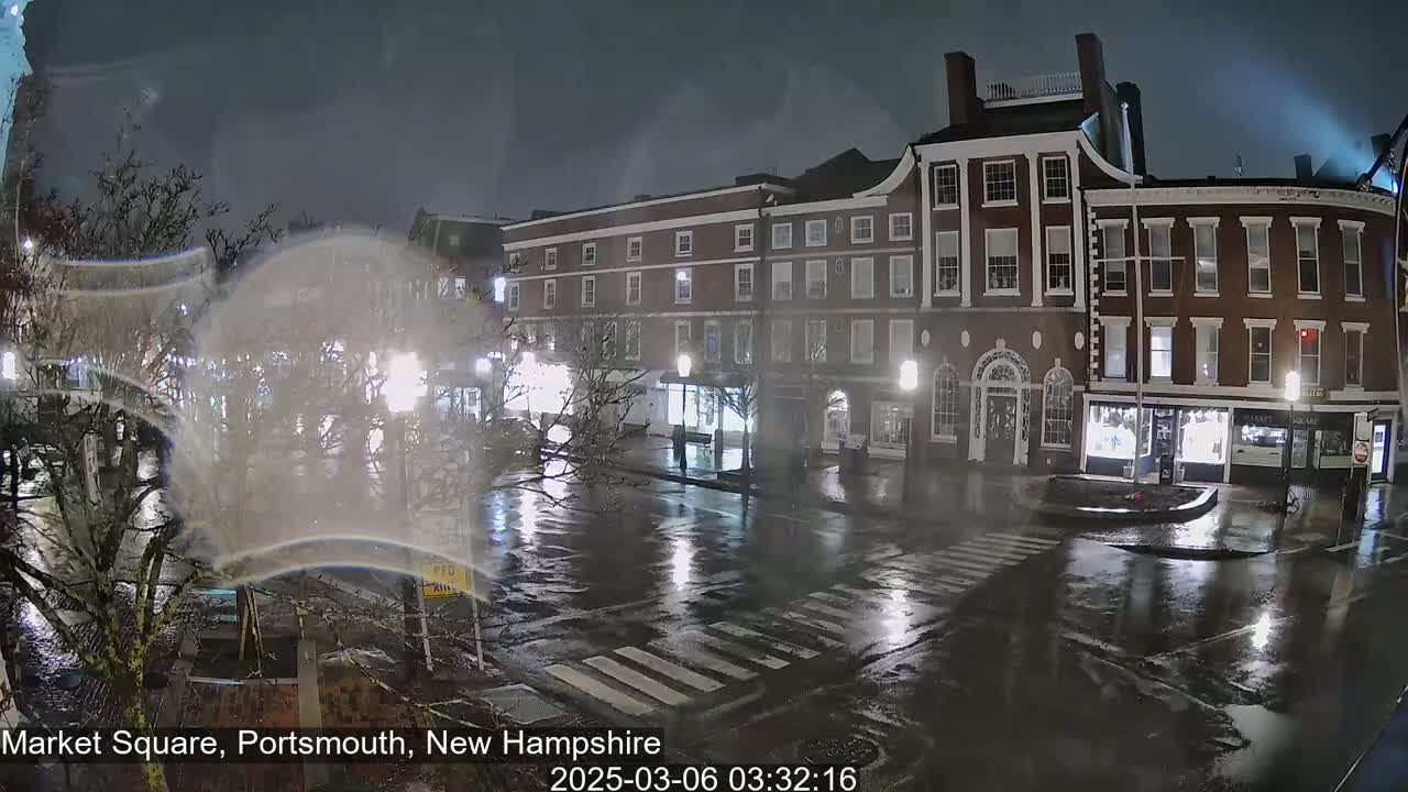 A nighttime view of a rain-slicked town square with several multi-story brick buildings under a dark sky.