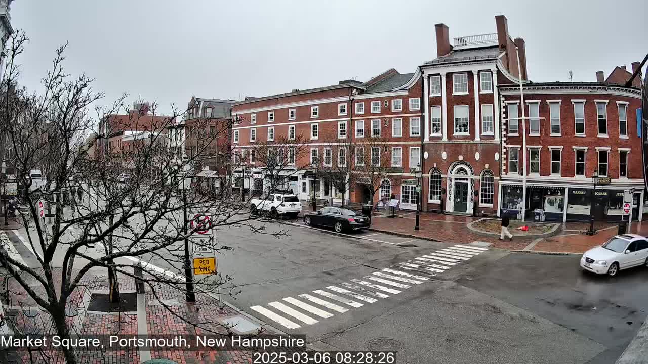 A street scene on an overcast day shows several brick buildings lining a square with cars parked and driving on a street that has a crosswalk.